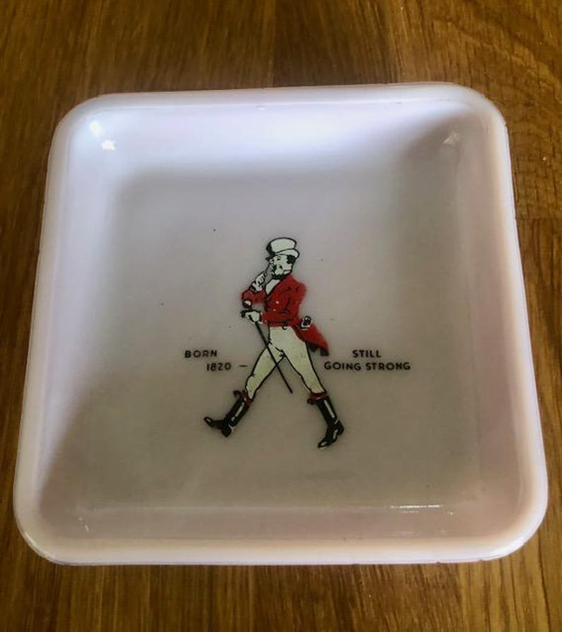May include: A small, square, pale pink dish with rounded corners. The center features a graphic of a man in a red coat and black boots, with the text "BORN 1820 - STILL GOING STRONG".