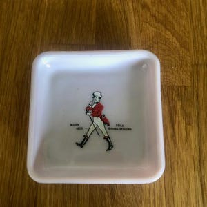 May include: Square, white ceramic tray with a red and black illustration of a man walking with a cane. Text on the tray reads "BORN 1820" and "STILL GOING STRONG". The tray is on a wooden surface.