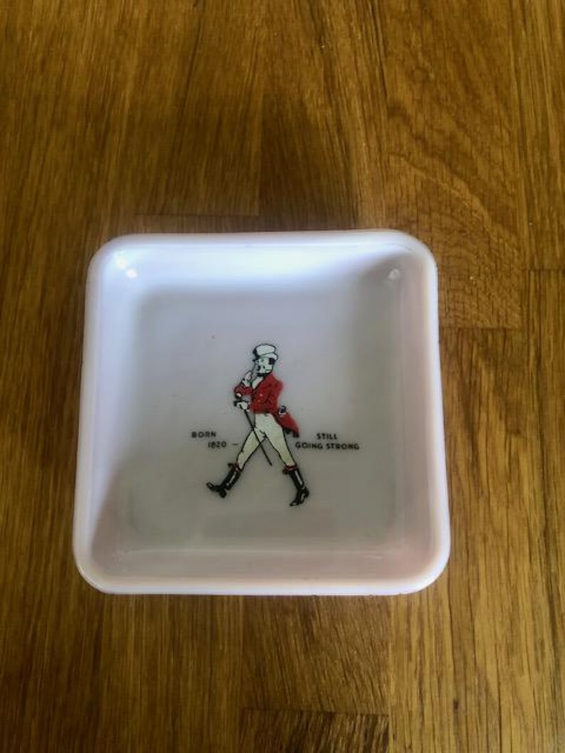 May include: Square, white ceramic tray with a red and black illustration of a man walking with a cane. Text on the tray reads "BORN 1820" and "STILL GOING STRONG". The tray is on a wooden surface.