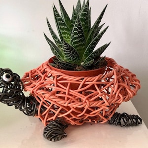 Puede incluir: Una maceta decorativa con forma de tortuga. El cuerpo está tejido en color coral, con cabeza y patas de color gris oscuro. Una planta suculenta se encuentra en el interior. La tortuga tiene un ojo blanco. La maceta mide unos 25 cm de largo.
