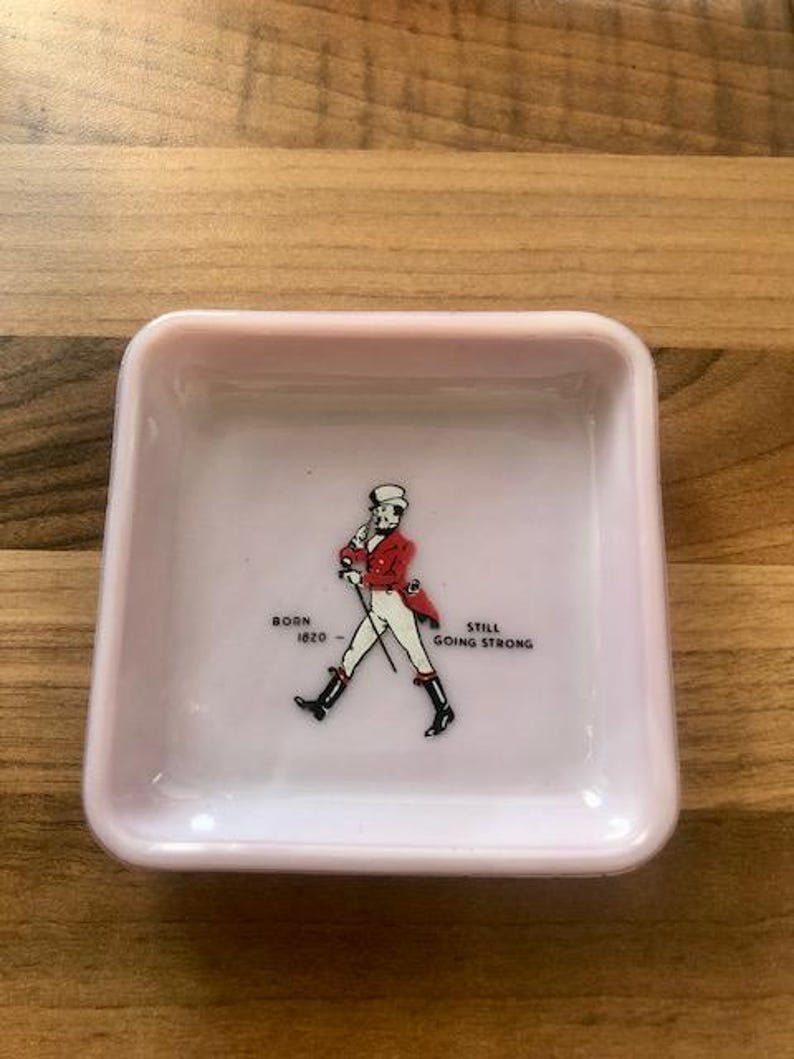 May include: A small, square, light pink dish. The center features a graphic of a man in a red coat and black boots, with the text "BORN 1820 - STILL GOING STRONG". The dish has a glossy finish and is likely ceramic.