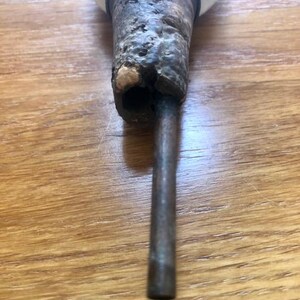 K&ouml;nnte beinhalten: Nahaufnahme eines Vintage-Flaschenverschlusses. Der Verschluss hat einen verwitterten, strukturierten Holzgriff mit dunklem, gealtertem Aussehen. Eine Metallstange erstreckt sich vom Griff, und eine wei&szlig;e, abgerundete Oberseite ist sichtbar.
