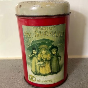 May include: Vintage red tin with a cream-colored lid. The tin features a label with the text "Chocolat Ph. Suchard" and an illustration of three children under an umbrella. The word "Neuchatel" is also visible.