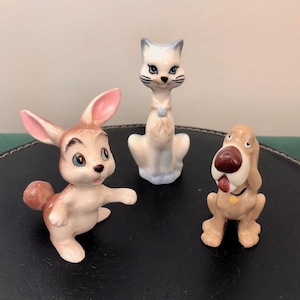 May include: Three ceramic figurines of a cat, rabbit, and dog. The cat is white with blue accents and has a yellow collar. The rabbit is brown and white with pink ears. The dog is brown with a red nose and a yellow collar.