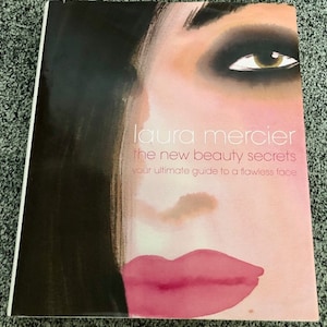 May include: A book cover with a close-up of a woman's face with heavy eye makeup and pink lips. The title of the book is "laura mercier the new beauty secrets your ultimate guide to a flawless face".