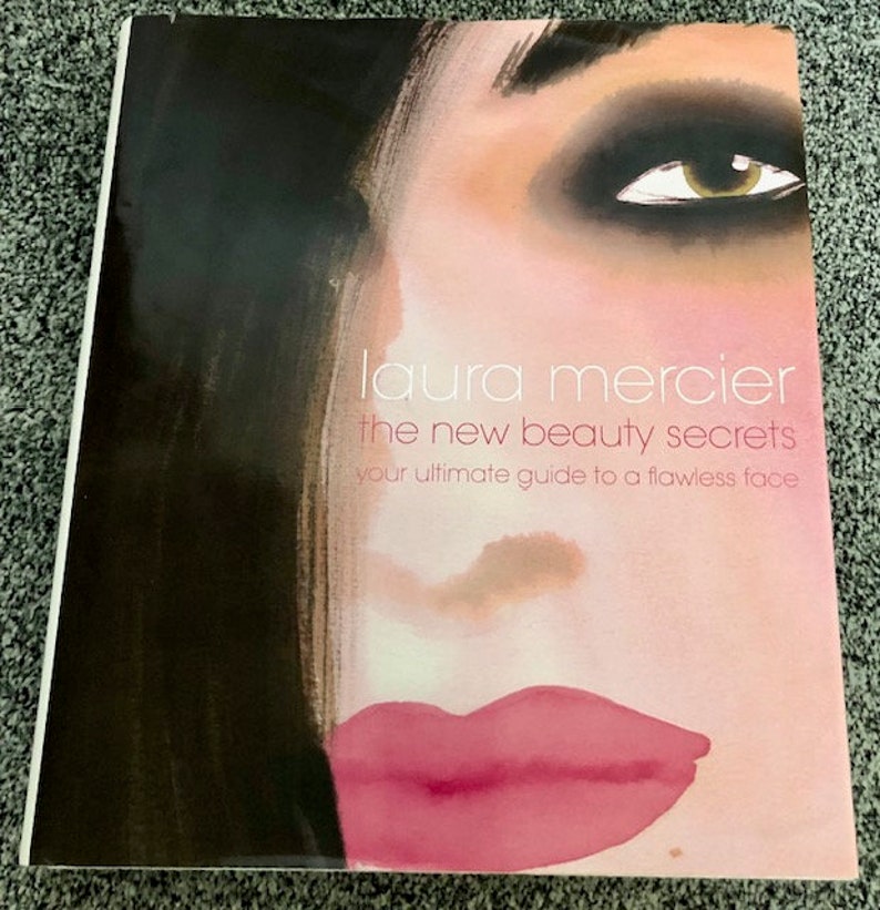 May include: A book cover with a close-up of a woman's face with heavy eye makeup and pink lips. The title of the book is "laura mercier the new beauty secrets your ultimate guide to a flawless face".