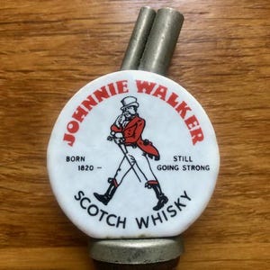 K&ouml;nnte beinhalten: Johnnie Walker Scotch Whisky Flaschenverschluss mit wei&szlig;em und rotem Logo. Das Logo zeigt eine Figur beim Gehen und den Text "Born 1820 - Still Going Strong". Er hat zwei Metallausl&auml;ufe.