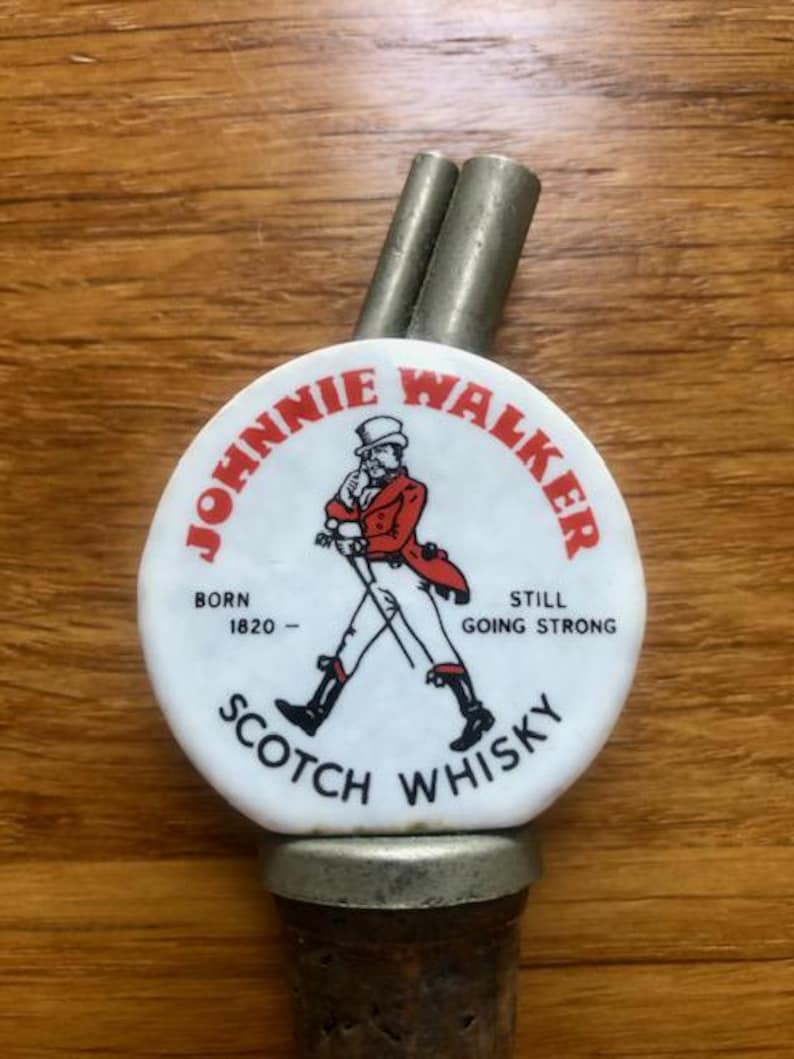 K&ouml;nnte beinhalten: Johnnie Walker Scotch Whisky Flaschenverschluss mit wei&szlig;em und rotem Logo. Das Logo zeigt eine Figur beim Gehen und den Text "Born 1820 - Still Going Strong". Er hat zwei Metallausl&auml;ufe.