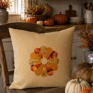 Könnte beinhalten: Ein quadratisches Dekokissen mit gelb-weißem Vichy-Karo-Muster. In der Mitte des Kissens befindet sich ein kreisförmiges Applikationsdesign, das aus abwechselnden Stoffblütenblättern in Unigelb, Kürbisdruck und gemustertem orangefarbenem Stoff besteht.