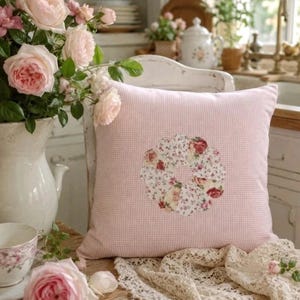 Könnte beinhalten: Ein quadratisches Zierkissen mit einem hellrosa-weißen Vichy-Karo-Muster und einer zentralen, kreisförmigen Applikation aus floralen Stoffstücken in Rosa-, Rot- und Grüntönen, die in einem gebogten, blumenartigen Design angeordnet sind.