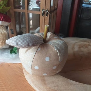 Könnte beinhalten: Ein beigefarbener Stoffapfel mit weißen Punkten und einem braunen Stiel. Der Apfel liegt auf einer Holzoberfläche.