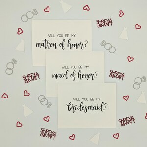May include: Three white cards with black calligraphy text asking "Will you be my matron of honor?", "Will you be my maid of honor?", and "Will you be my bridesmaid?" Scattered around are red hearts, silver rings, and white dress-shaped confetti.