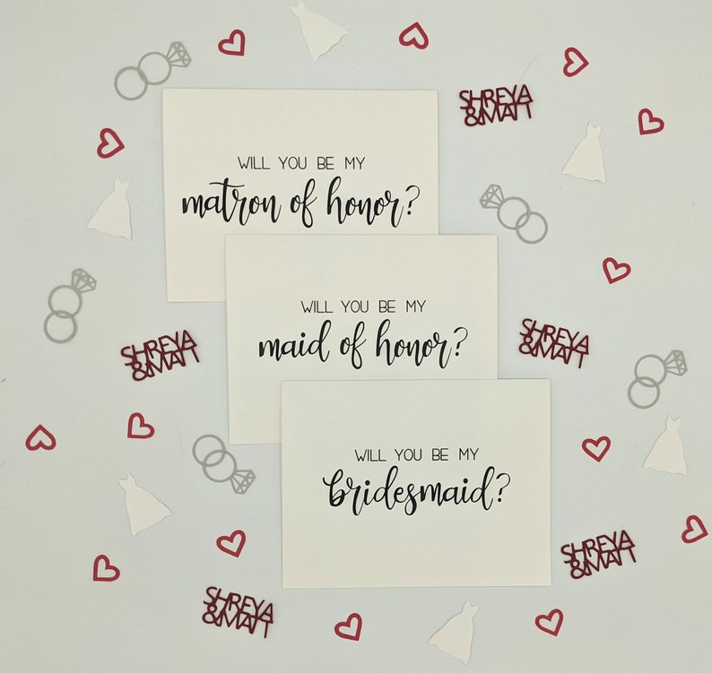 May include: Three white cards with black calligraphy text asking "Will you be my matron of honor?", "Will you be my maid of honor?", and "Will you be my bridesmaid?" Scattered around are red hearts, silver rings, and white dress-shaped confetti.