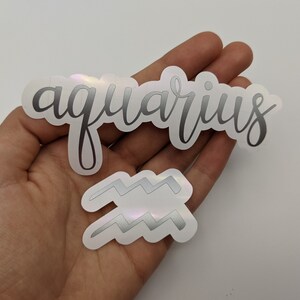May include: Two holographic stickers featuring the word aquarius in a cursive script font and a separate sticker depicting the Aquarius zodiac symbol, which consists of two parallel horizontal zigzag lines, both with a metallic silver finish.