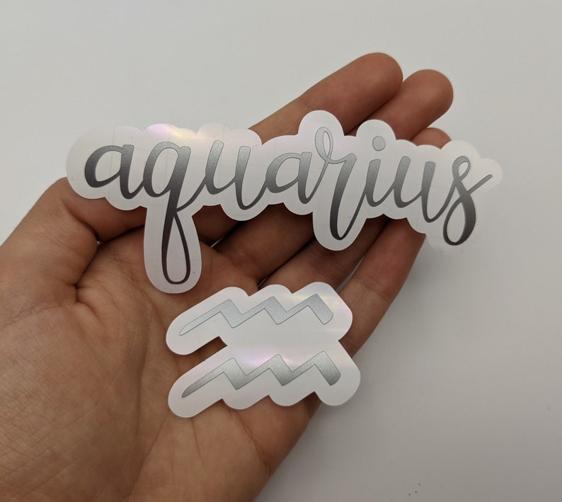 May include: Two holographic stickers featuring the word aquarius in a cursive script font and a separate sticker depicting the Aquarius zodiac symbol, which consists of two parallel horizontal zigzag lines, both with a metallic silver finish.