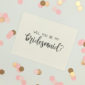 May include: A white card with the handwritten text "WILL YOU BE MY bridesmaid?" in black script. The card is surrounded by pink and gold confetti on a white background. A wedding invitation or proposal.