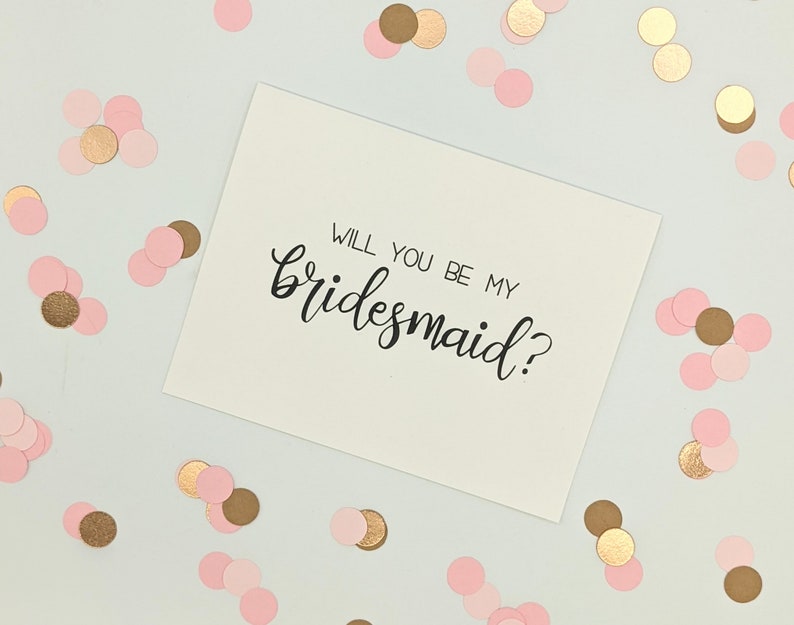 May include: A white card with the handwritten text "WILL YOU BE MY bridesmaid?" in black script. The card is surrounded by pink and gold confetti on a white background. A wedding invitation or proposal.