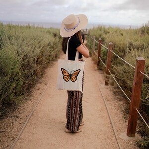 Puede incluir: Una persona con un sombrero de paja de ala ancha y una blusa negra lleva una bolsa de tela blanca con un dise&ntilde;o de mariposa naranja. La persona est&aacute; de pie en un camino de tierra con una valla de madera en el lado derecho.