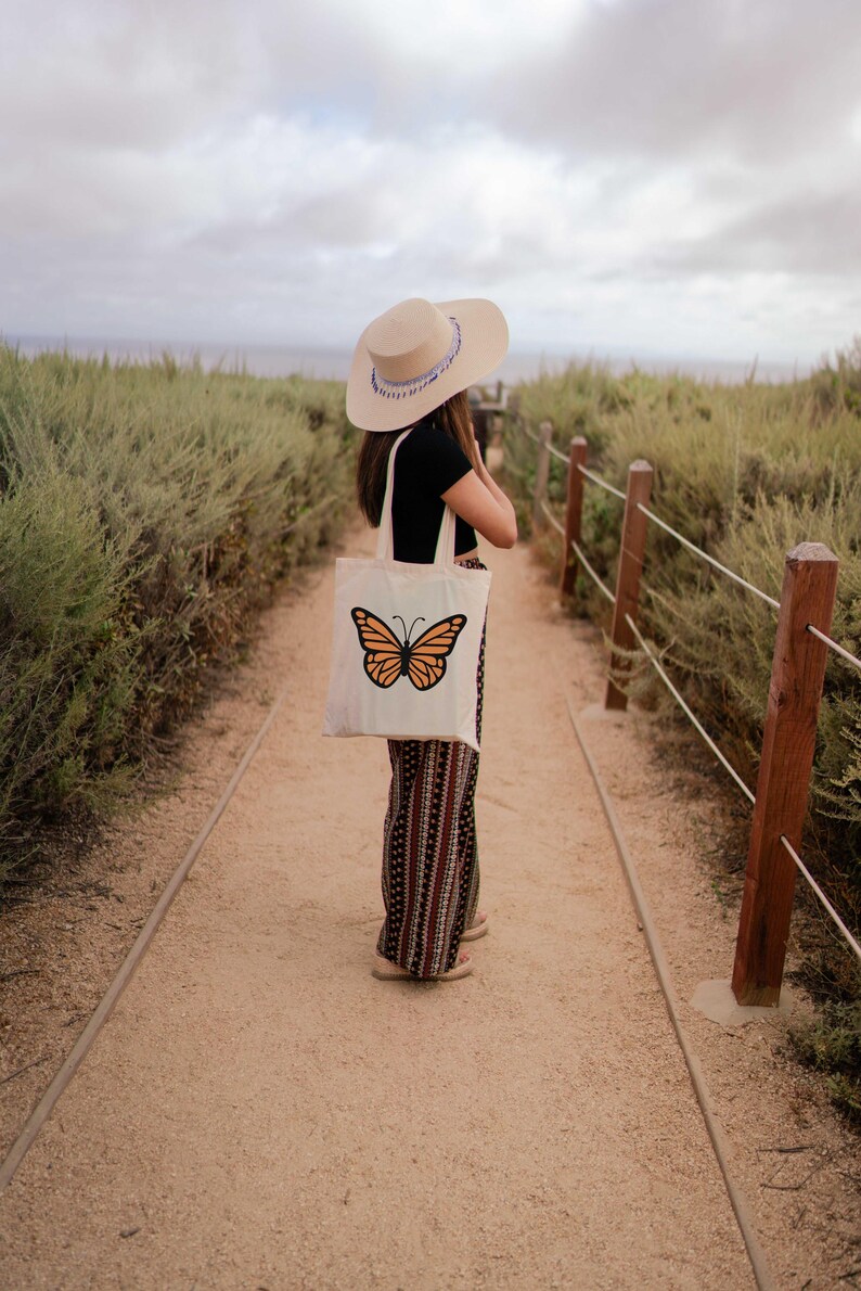 Puede incluir: Una persona con un sombrero de paja de ala ancha y una blusa negra lleva una bolsa de tela blanca con un dise&ntilde;o de mariposa naranja. La persona est&aacute; de pie en un camino de tierra con una valla de madera en el lado derecho.