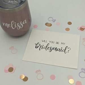 May include: A rose gold tumbler with the name "melissa" in white script. A card with the text "WILL YOU BE MY bridesmaid?" is surrounded by pink, gold, and silver confetti in the shape of rings and circles.