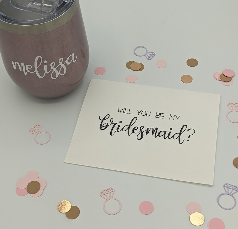 May include: A rose gold tumbler with the name "melissa" in white script. A card with the text "WILL YOU BE MY bridesmaid?" is surrounded by pink, gold, and silver confetti in the shape of rings and circles.