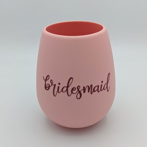 May include: A stemless, blush pink wine glass with the word "bridesmaid" in a dark brown script. The interior of the glass is a darker shade of pink. The glass has a smooth, matte finish.