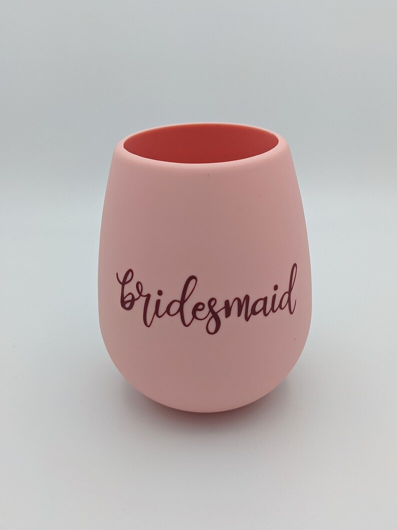 May include: A stemless, blush pink wine glass with the word "bridesmaid" in a dark brown script. The interior of the glass is a darker shade of pink. The glass has a smooth, matte finish.