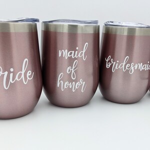 May include: Four rose gold stemless wine tumblers with silver rims and lids. The tumblers are labeled with white script text: "bride", "maid of honor", and two "bridesmaid" labels. Ideal for bridal parties.