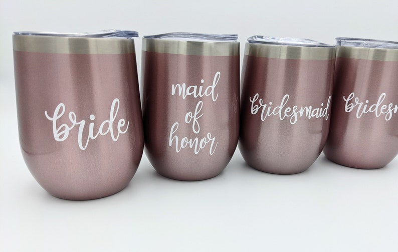 May include: Four rose gold stemless wine tumblers with silver rims and lids. The tumblers are labeled with white script text: "bride", "maid of honor", and two "bridesmaid" labels. Ideal for bridal parties.