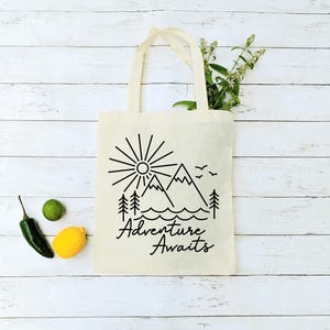 May include: A white canvas tote bag with black line art of mountains, a sun, and trees. The text "Adventure Awaits" is printed in black below the image.