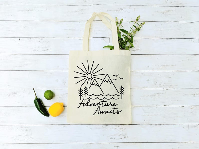 May include: A white canvas tote bag with black line art of mountains, a sun, and trees. The text "Adventure Awaits" is printed in black below the image.