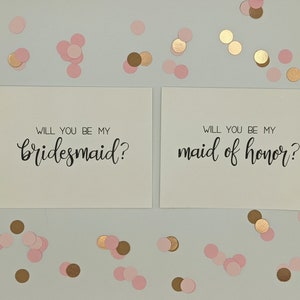 May include: Two white cards with black calligraphy text that reads "Will you be my bridesmaid?" and "Will you be my maid of honor?" Scattered around are pink and gold confetti. A wedding or bridal party theme.