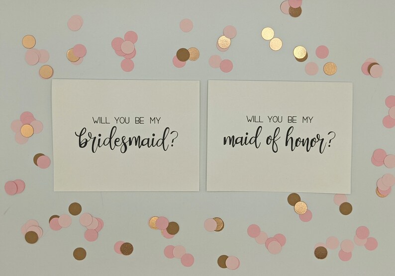 May include: Two white cards with black calligraphy text that reads "Will you be my bridesmaid?" and "Will you be my maid of honor?" Scattered around are pink and gold confetti. A wedding or bridal party theme.
