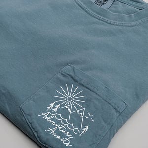 May include: A light blue t-shirt with a pocket featuring a white graphic of mountains, a sun, and trees with the text "Adventure Awaits".