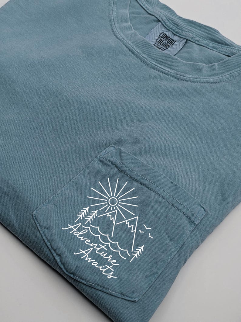May include: A light blue t-shirt with a pocket featuring a white graphic of mountains, a sun, and trees with the text "Adventure Awaits".