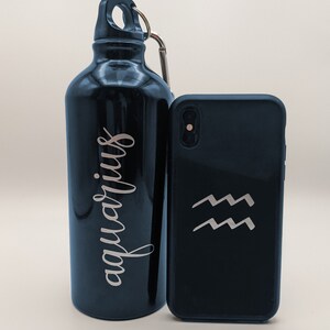 May include: A glossy black metal water bottle with a silver carabiner and the word "aquarius" written in a cursive script, positioned next to a black smartphone case featuring a silver Aquarius zodiac symbol consisting of two wavy lines.
