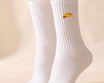 May include: A pair of white ribbed crew socks featuring a small embroidered graphic of a yellow duckling resting on the outer side of each sock. The socks are made of a knit fabric and extend to the mid-calf, showcasing a clean and simple design.