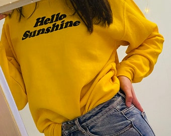 May include: A yellow sweatshirt with the text "Hello Sunshine" in black lettering. The sweatshirt is worn over blue jeans.