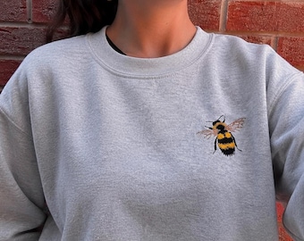 May include: A light gray crewneck sweatshirt featuring a small, detailed embroidered bumblebee on the left chest. The bee is depicted with yellow and black stripes, black legs, and translucent wings, creating a clear contrast against the fabric.