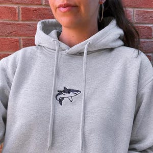 Könnte beinhalten: Ein hellgrauer Kapuzenpullover mit einer kleinen, gestickten Haigrafik in der Mitte der Brust. Der Hoodie verfügt über eine passende Kordel und eine Vordertasche und ist aus einem weichen, melierten Stoff mit langen Ärmeln gefertigt.