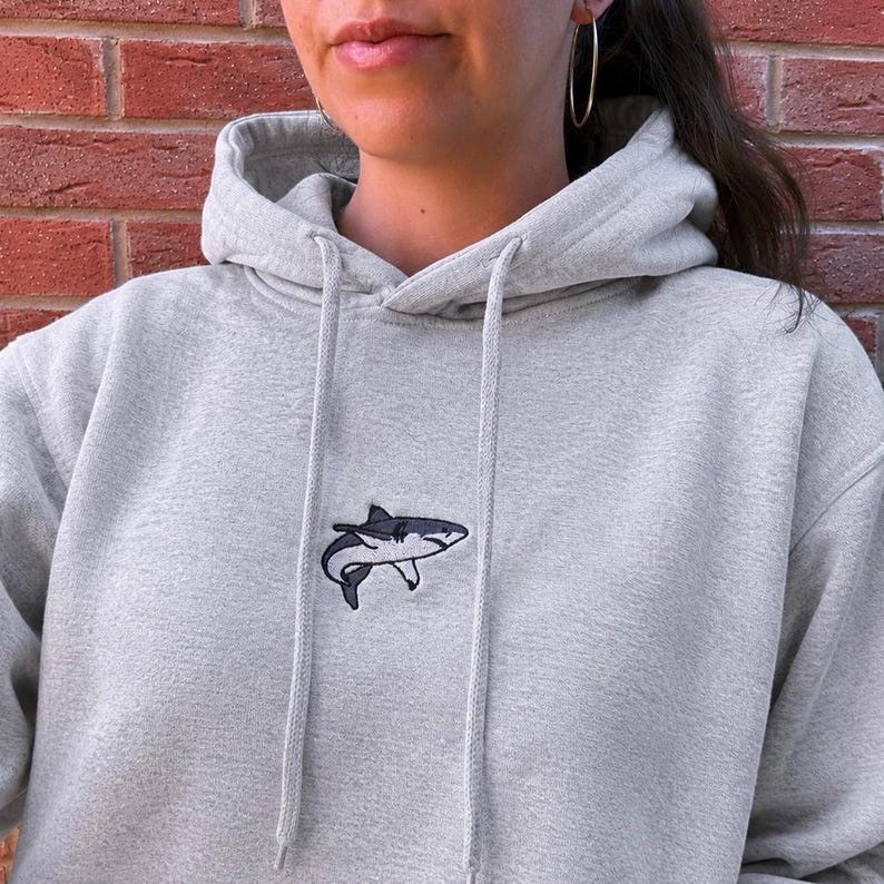 May include: A light grey hooded sweatshirt featuring a small, embroidered shark graphic centred on the chest. The hoodie includes a matching drawstring cord and a front pocket, crafted from a soft, heathered fabric with long sleeves.