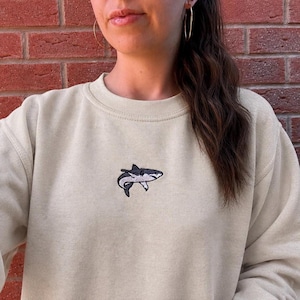 May include: A beige crewneck sweatshirt featuring a small, centered embroidered graphic of a grey and white shark. The garment has long sleeves and a ribbed neckline, providing a simple and clean look with the detailed shark design.
