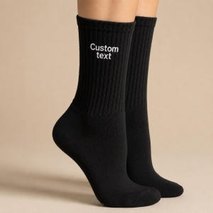 May include: A pair of black crew-length socks featuring a ribbed texture throughout the leg. The outer side of the left sock displays the words Custom text embroidered in white thread, positioned clearly on the upper portion of the ribbed cuff.