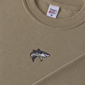 May include: A tan crewneck sweatshirt featuring a small embroidered shark graphic on the chest. The interior label reads Embroidery in the Attic and indicates size M. The fabric has a smooth texture with a ribbed collar and long sleeves.