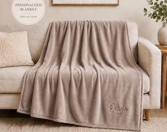 May include: A solid taupe-colored plush blanket draped over a sofa. The blanket features the embroidered name Olivia in a cursive script font, positioned above a small embroidered heart outline in the lower right corner of the fabric.