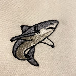 May include: An embroidered patch of a shark on a light beige fabric background. The shark is depicted in shades of dark grey, light grey, and white, featuring a black stitched outline, visible gills, and a curved body shape facing towards the right.