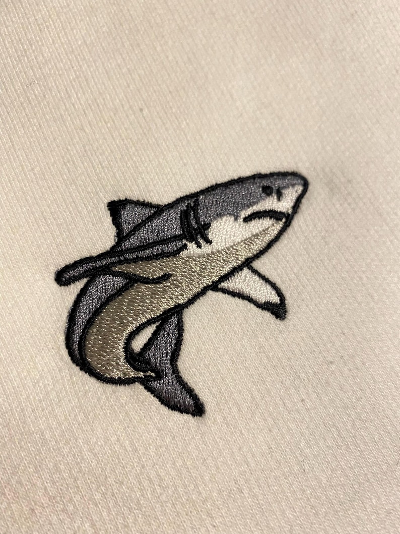 May include: An embroidered patch of a shark on a light beige fabric background. The shark is depicted in shades of dark grey, light grey, and white, featuring a black stitched outline, visible gills, and a curved body shape facing towards the right.