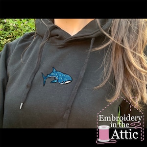 May include: Black hoodie with a blue embroidered whale shark on the front. The hoodie has a drawstring hood and a kangaroo pocket. The text "Embroidery in the Attic" is visible in the bottom right corner of the image.