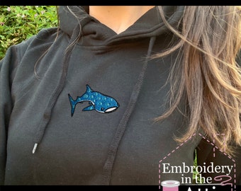 Può includere: Felpa con cappuccio nera con uno squalo balena ricamato in blu sul davanti. La felpa ha un cappuccio con coulisse e una tasca a marsupio. Il testo "Embroidery in the Attic" è visibile nell'angolo in basso a destra dell'immagine.