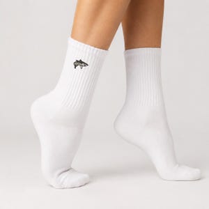 May include: A pair of white crew-length socks featuring a small, embroidered graphic of a grey shark on the outer side of the left sock. The socks have a ribbed texture at the ankle and are made of a solid white fabric with a clean, simple design.